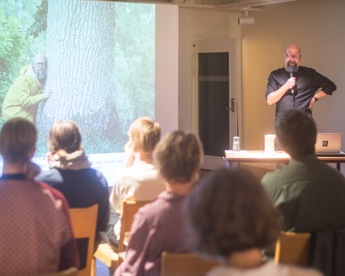 Man står framför en skärm och håller en presentation. På bilden syns samme man som lyssnar med örat mot ett träd.